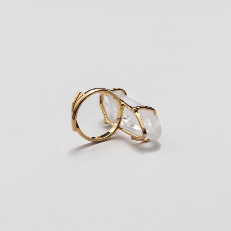 May include: A gold ring with a large, clear crystal. The crystal is rectangular and held in a gold frame. The ring band has a delicate, organic design. The overall style is elegant and minimalist, suitable for everyday wear.