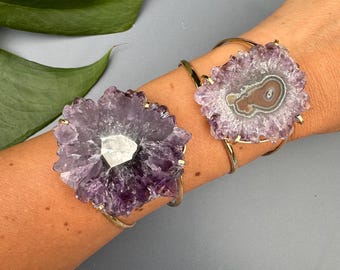 May include: Two gold-coloured bracelets, each with a large, purple amethyst crystal. The crystals have a unique, flower-like shape with a central white quartz accent. The bracelets are displayed on an arm against a grey background.