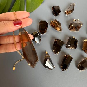May include: A gold chain necklace with a large, faceted, smoky quartz pendant. Several loose smoky quartz crystals with gold accents are scattered around the necklace on a gray surface. The crystals vary in size and shape.
