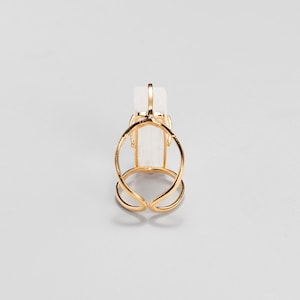 May include: A gold-toned ring featuring an open, double-band design that curves around a central, vertically oriented, translucent white rectangular crystal stone. The metal band is polished and smooth, creating a continuous frame around the stone.