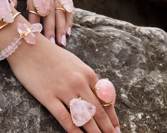 May include: A collection of gold-toned rings and a bracelet featuring pale pink rose quartz gemstones. The rings showcase various shapes including oval, triangular, and teardrop cuts, all set in gold metal bands. A beaded bracelet features a central double-terminated rose quartz crystal point.