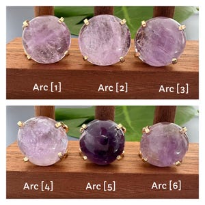 May include: Six amethyst rings with gold settings displayed on a wooden surface. The rings are arranged in two rows of three. The amethyst stones are various shades of purple.