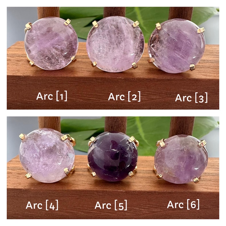 May include: Six amethyst rings with gold settings displayed on a wooden surface. The rings are arranged in two rows of three. The amethyst stones are various shades of purple.