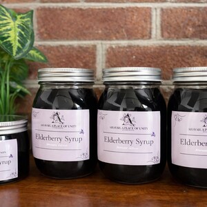 Peut inclure: Quatre pots en verre de sirop de sureau avec des couvercles argentés sont disposés sur une surface en bois. Chaque pot porte une étiquette indiquant "Elderberry Syrup". Une petite plante aux feuilles vertes et jaunes se trouve à gauche des pots. L'arrière-plan est un mur de briques.
