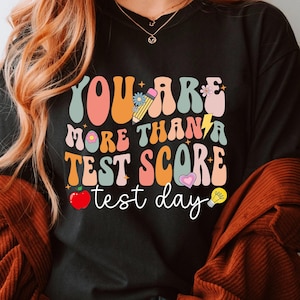 May include: Black t-shirt with a colorful retro graphic that says "You are more than a test score test day". The graphic includes a pencil, lightning bolt, lightbulb, apple, and heart.