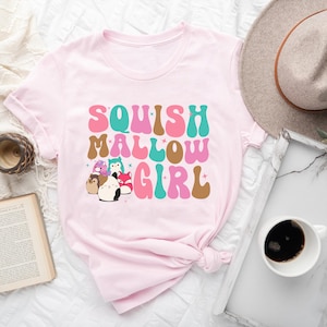 Puede incluir: Camiseta rosa con el texto "SQUISH MALLOW GIRL" en una fuente retro. El texto está en tonos rosa, marrón, turquesa y verde. Hay pequeños animales de dibujos animados en la esquina inferior izquierda de la camiseta.