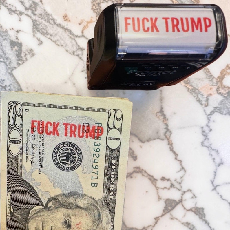 May include: A black self-inking rubber stamp with the red text FUCK TRUMP displayed on the top label, positioned next to a twenty-dollar bill that has been stamped with the same red text FUCK TRUMP across the center of the currency note.