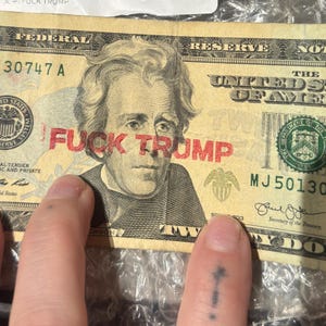 May include: A twenty-dollar bill featuring a portrait of Andrew Jackson with the red stamped text FUCK TRUMP across the center. The bill includes standard federal reserve markings, serial numbers, and the United States of America text.