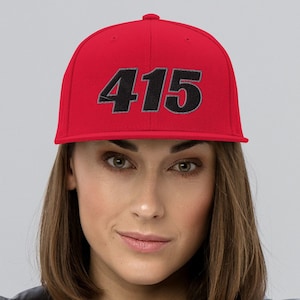 Może przedstawiać: Czerwona czapka bejsbolowa z numerem "415" wyhaftowanym w kolorze czarnym i szarym. Czapka ma płaski daszek i strukturalny krój. Tło jest jednolite, jasnoszare.