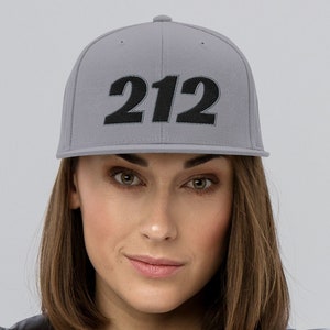 May include: A grey baseball cap featuring the number "212" embroidered in black. The cap is worn by a person wearing a grey jacket over a black and white striped top. The background is a plain light grey.