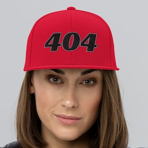 Könnte beinhalten: Rote Baseballkappe mit der schwarzen Aufschrift "404". Die Kappe wird von einer Person getragen, die eine graue Jacke über einem schwarz-weiß gestreiften Oberteil trägt. Die Kappe hat einen flachen Schirm.