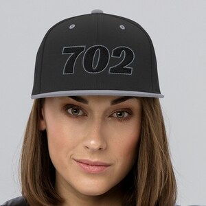 Może przedstawiać: Czarna czapka z daszkiem z płaskim daszkiem, z wyszytym numerem "702" w kolorze szarym. Czapka ma szary daszek i szary guzik na górze. Osoba na zdjęciu ma na sobie szarą kurtkę.