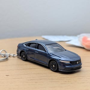 May include: A dark blue metallic saloon toy car keyring featuring a detailed front grille with a small Honda emblem, black wheels, and a silver metal keyring attached to the rear. The miniature vehicle has a sleek body design with tinted windows.