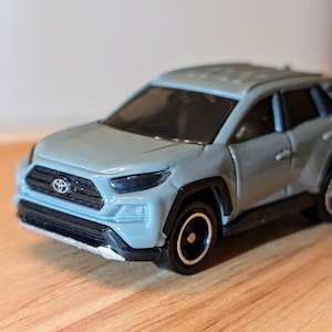 Pode incluir: Um SUV de brinquedo em metal fundido na cor cinza claro, com grade frontal preta e emblema da Toyota, janelas escuras, arcos de roda pretos e rodas pretas com aros prateados. O veículo possui um design moderno com para-choque dianteiro prateado.