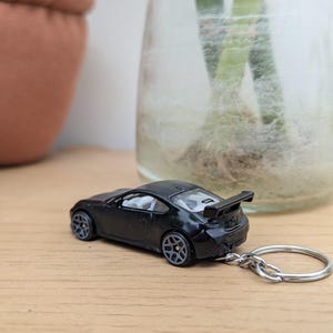 May include: Black toy car keychain with a silver key ring. The car has a rear spoiler and is sitting on a wooden surface.