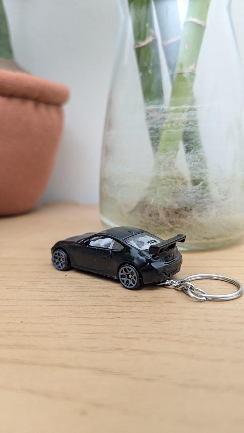 May include: Black toy car keychain with a silver key ring. The car has a rear spoiler and is sitting on a wooden surface.