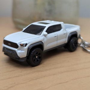 May include: White toy truck keychain with black wheels. The truck has a black grille and black accents.