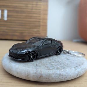 May include: A black toy car keychain with a silver key ring. The car is a sports car with a sleek design. The car is sitting on a gray stone.