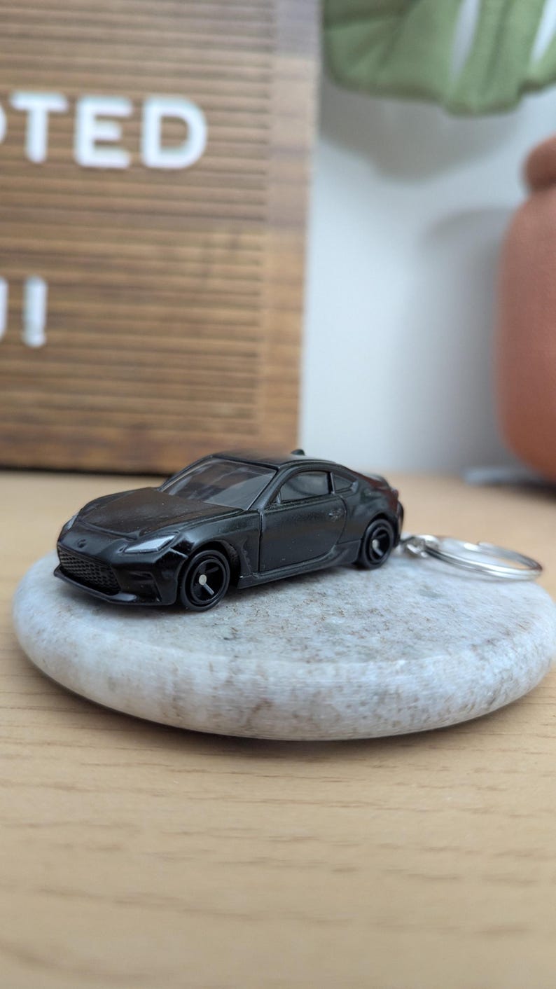 May include: A black toy car keychain with a silver key ring. The car is a sports car with a sleek design. The car is sitting on a gray stone.
