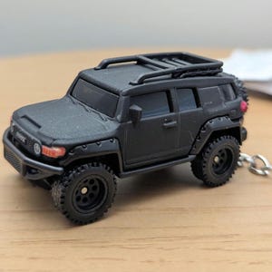 Może przedstawiać: Czarny model odlewu ciśnieniowego SUV-a Toyota FJ Cruiser. Miniaturowy pojazd posiada bagażnik dachowy, wytrzymałe opony i brelok do kluczy. Reflektory i tylne światła mają czerwone i pomarańczowe akcenty. To przedmiot kolekcjonerski.
