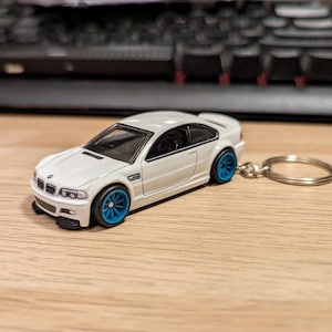 May include: A white BMW M3 model car keychain with blue wheels. The miniature car has a detailed design, including a spoiler and black accents. The keychain is attached to a silver metal ring.