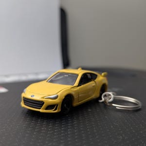 May include: A yellow die-cast model car keychain. The miniature sports car has black wheels and a silver key ring. The car's design includes a front grill, headlights, and a rear spoiler. The keychain is a miniature replica of a sports car.