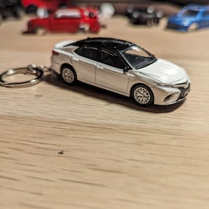 Könnte beinhalten: Ein weißes Spielzeugauto mit schwarzem Dach und einem silbernen Schlüsselanhänger. Das Auto steht auf einer Holzoberfläche.