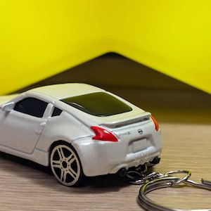 May include: A white toy car keychain with a silver key ring. The car is a Nissan 370Z and has a black interior and black wheels.