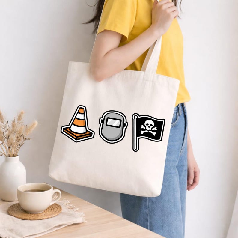 May include: Off-white tote bag featuring three cartoon graphics: an orange and white traffic cone, a gray welding mask, and a black pirate flag with a skull and crossbones. The bag is held by a person wearing a yellow shirt and blue jeans.