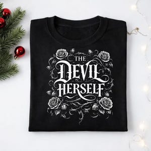 Puede incluir: Sudadera negra con el texto "THE DEVIL HERSELF" en una fuente decorativa, rodeada de rosas blancas y vides. El diseño está centrado en la parte delantera de la prenda. La sudadera es de cuello redondo.