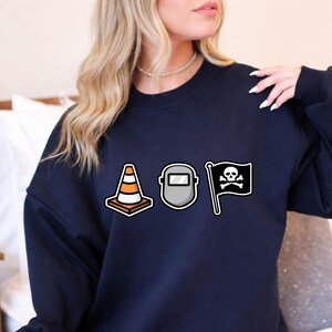 May include: Navy blue sweatshirt featuring three graphic designs: a traffic cone, a welding mask, and a pirate flag with a skull and crossbones. The sweatshirt is a solid color and has a crew neck.