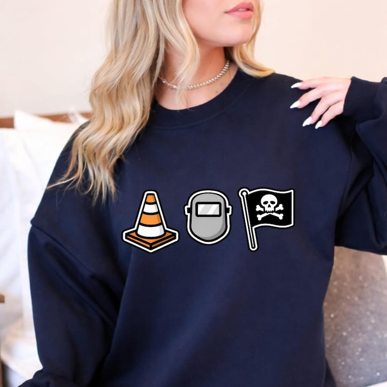 May include: Navy blue sweatshirt featuring three graphic designs: a traffic cone, a welding mask, and a pirate flag with a skull and crossbones. The sweatshirt is a solid color and has a crew neck.