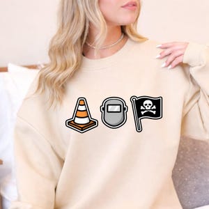 May include: Beige sweatshirt featuring three graphic icons: a traffic cone, a welding mask, and a pirate flag with a skull and crossbones. The sweatshirt is a neutral color and has a crew neck.