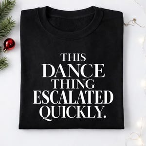 Puede incluir: Sudadera negra con cuello redondo con el texto blanco: "THIS DANCE THING ESCALATED QUICKLY". La sudadera está doblada sobre una superficie blanca, con decoraciones festivas en el fondo.