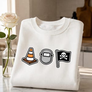 May include: White sweatshirt featuring three cartoon graphics: an orange and white traffic cone, a gray welding mask, and a black flag with a skull and crossbones. The sweatshirt is folded on a marble surface.