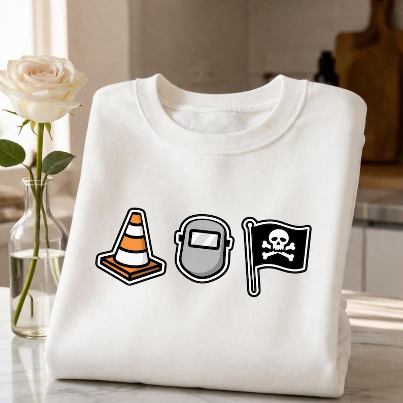 May include: White sweatshirt featuring three cartoon graphics: an orange and white traffic cone, a gray welding mask, and a black flag with a skull and crossbones. The sweatshirt is folded on a marble surface.
