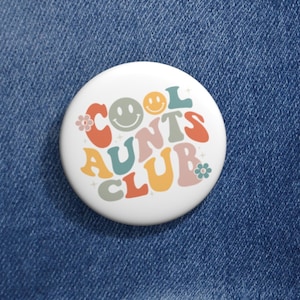 May include: A white button with a colorful retro design that says "Cool Aunts Club" with smiley faces and flowers.