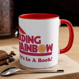 May include: White ceramic mug with a red rim and handle. The mug features a red and yellow logo that says "Reading Rainbow It's In A Book!"