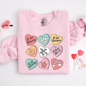 May include: Pink sweatshirt with a graphic of colorful hearts with positive affirmations like "I am strong", "I choose joy", and "I am worthy".