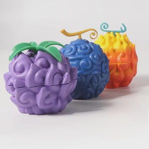 Op de afbeelding: Drie kleurrijke, gestileerde figuren in de vorm van fruit. De eerste is paars met groene bladeren, de tweede is blauw met een gouden steel en de derde is geel en oranje. Elk heeft een uniek ontwerp en textuur, wat een fantasiethema suggereert.