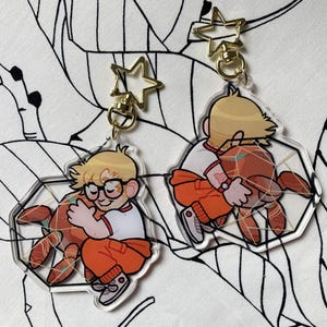 May include: Two clear acrylic keychains featuring a cartoon character with blonde hair and glasses wearing an orange outfit, embracing a large orange spider-like creature. Each keychain has a gold-colored star-shaped clasp at the top.