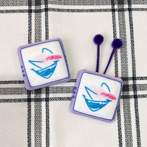 Pode incluir: Dois brincos quadrados com borda lavanda e centro branco com um rosto de desenho animado com boca azul, bochechas rosa e sobrancelha curva. Um tem antenas roxas.