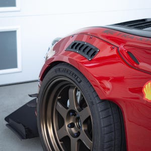 May include: A red car wing with a black vented panel and a wide-body kit. The vehicle features a bronze six-spoke wheel with a Michelin 265/35 ZR18 tyre. A black front splitter is visible, and the text TRA-KYOTO is printed on the bonnet.
