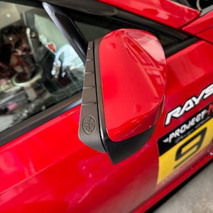 Könnte beinhalten: Ein leuchtend roter Auto-Seitenspiegel mit einer mattschwarzen Zierleiste und einem kreisförmigen Emblem an der Seite. Die Fahrzeugkarosserie zeigt teilweise weiße und gelbe Aufkleber mit dem Text RAYS und PROJECT M auf dem Seitenteil.