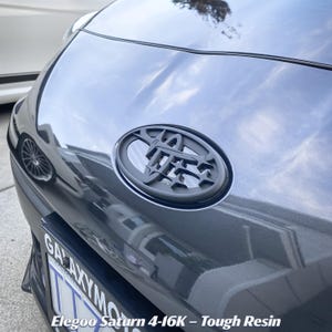 Pode incluir: Um emblema de carro personalizado em preto fosco com um design estilizado em forma de estrela montado no capô de um veículo cinza escuro. O emblema tem um padrão geométrico. Abaixo do emblema, uma moldura de placa exibe Elegoo Saturn 4-16K.