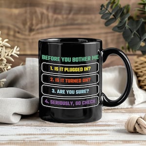 Könnte beinhalten: Schwarze Keramik-Kaffeetasse mit gebogenem Henkel. Die Tasse zeigt den Text "BEFORE YOU BOTHER ME" und eine Liste von Fragen in Weiß, Gelb, Rot und Lila. Die Fragen lauten: "1. IS IT PLUGGED IN?", "2. IS IT TURNED ON?", "3. ARE YOU SURE?" und "4. SERIOUSLY, GO CHECK".