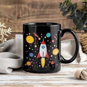 Peut inclure: Mug en céramique noire avec un motif spatial coloré. Une fusée blanche et rouge est entourée de planètes, d'étoiles et d'un soleil. La tasse a une anse noire et une finition brillante, idéale pour le café ou le thé.