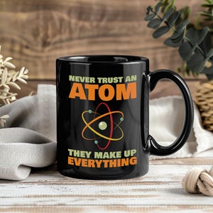 May include: Black ceramic mug with the text "NEVER TRUST AN ATOM THEY MAKE UP EVERYTHING" in orange and white. The mug features an atom graphic. The mug has a black handle and is on a wooden surface.