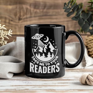 May include: Black ceramic mug with a white graphic of a UFO beaming down on mountains, trees, and the text "Take Me To Your Readers." The mug has a curved handle and is suitable for hot or cold beverages.