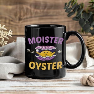 May include: Black ceramic mug with the text "Moister Than An Oyster" in purple and yellow. The mug features a cartoon oyster with a smiling face. The mug has a black handle and a glossy finish.
